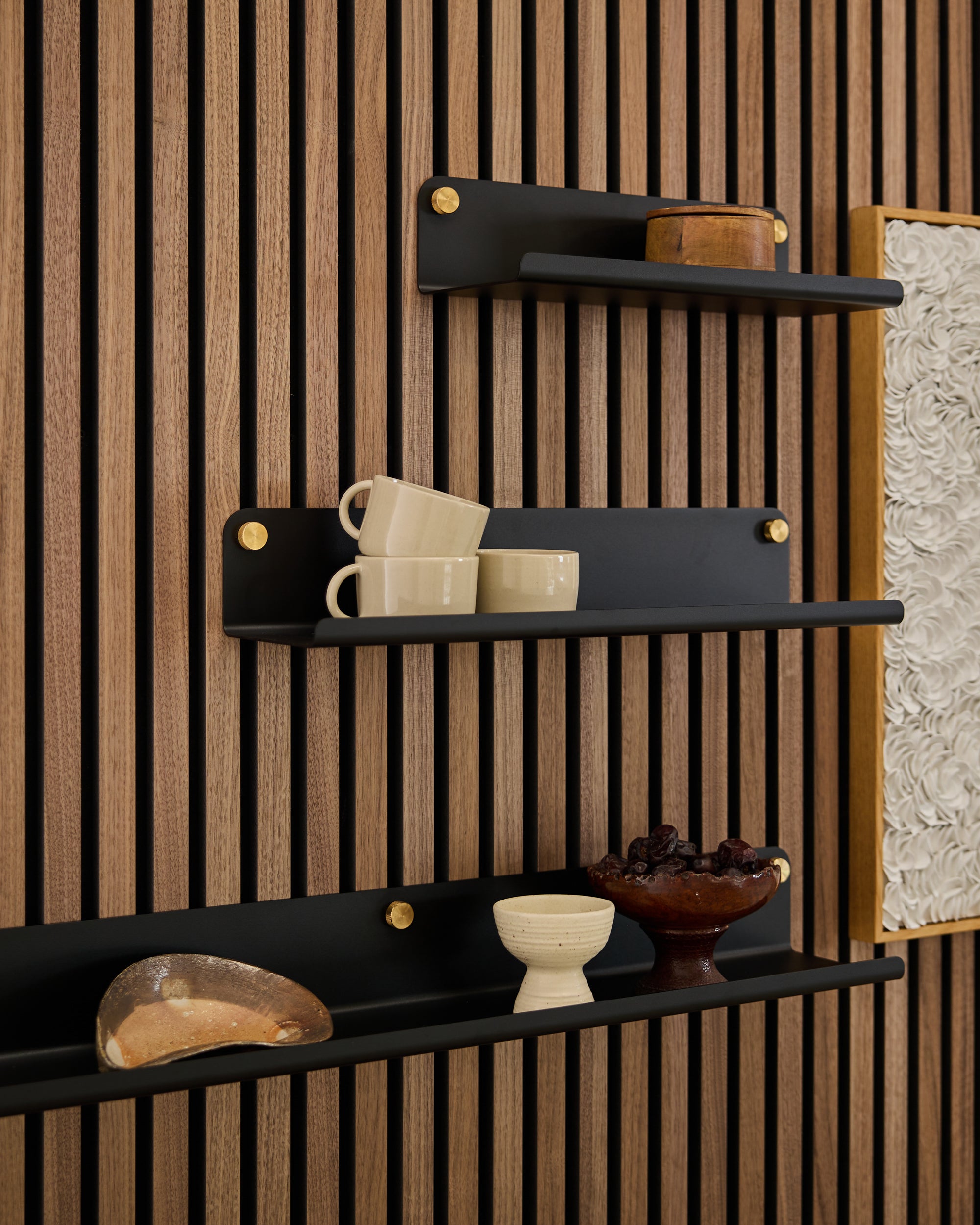 Three black wall shelves mounted on a Nordic wood Akupanel slatwall display cups, bowls, and decor; textured white acoustic art panel partially visible at right.